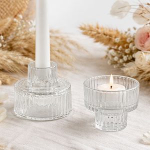 Szklane świeczniki w kształcie cylindrów, jeden z białą świecą, a drugi z płonącą świeczką; otoczone suszonymi kwiatami.