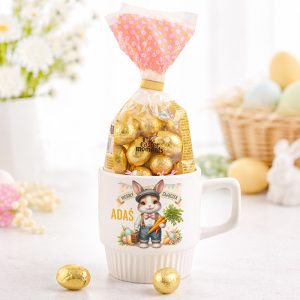 Kubek z rysunkiem zająca, wypełniony czekoladowymi jajkami, z opakowaniem na górze i złotymi jajkami obok.