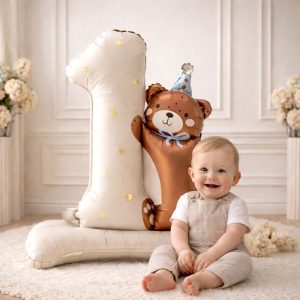 Mały chłopiec w szarej odzieży siedzi obok dużego balonu w kształcie liczby jeden z pluszowym misiem, w otoczeniu kwiatów.