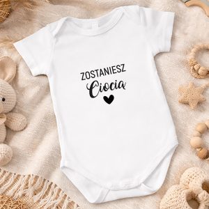 Biały body niemowlęce z napisem "Zostaniesz Ciocia" oraz serduszkiem. Otoczone pluszakami i naturalnymi detalami.