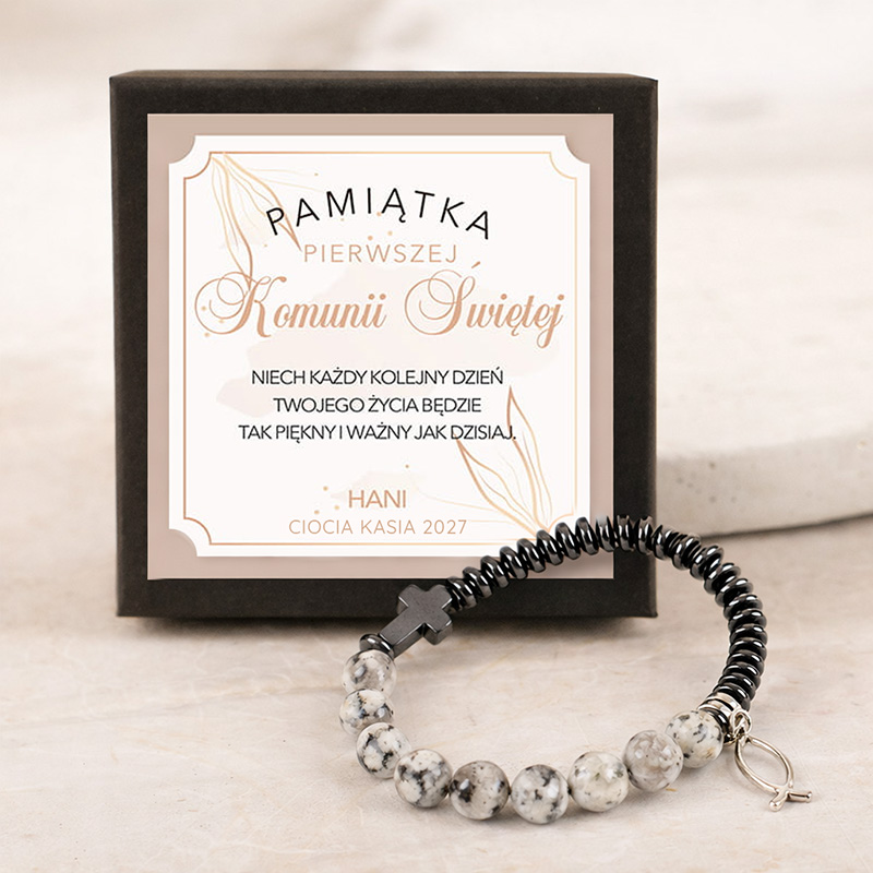 Bransoletka z czarnymi i białymi koralikami leży obok eleganckiego pudełka z napisem "Pamiątka Pierwszej Komunii Świętej".