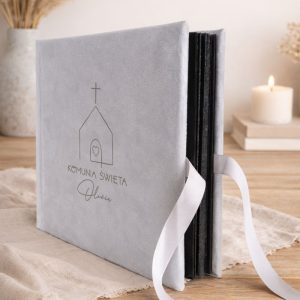 Szary album na zdjęcia z napisem "Komunia Święta" i rysunkiem kościoła, związany białą wstążką, na drewnianym stole.