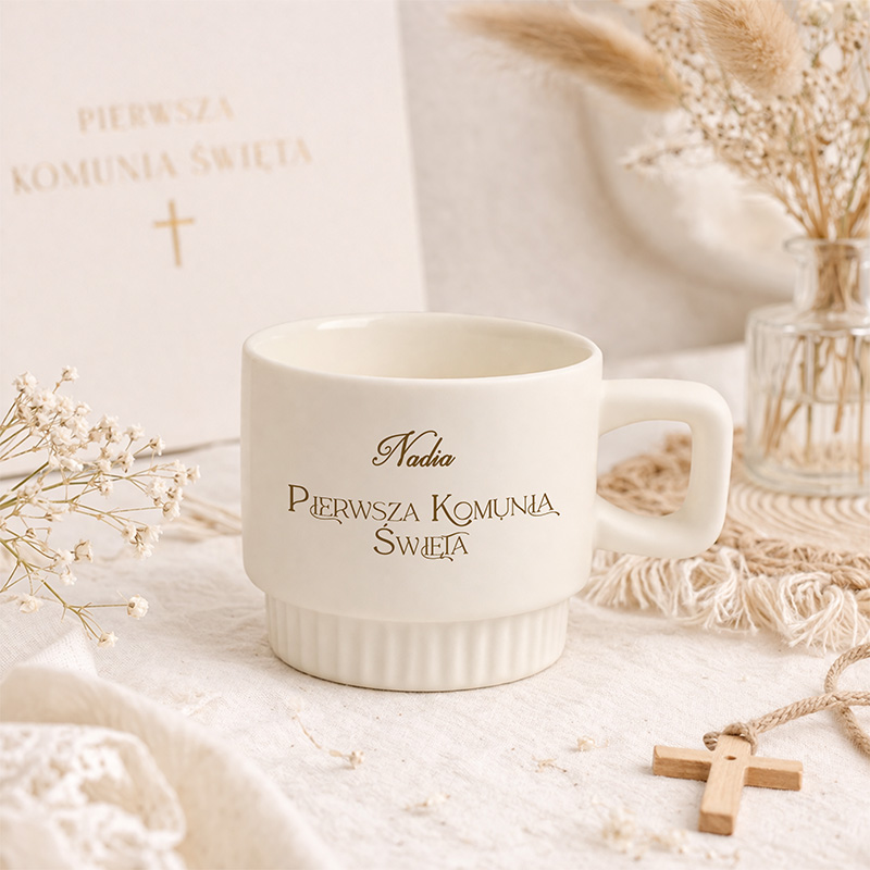 Biały kubek z napisem "Nadia Pierwsza Komunia Święta" na tle jasnego materiału i dekoracji.