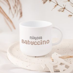 Biały kubek z napisem "nikola Babyccino" na białym talerzu, otoczony suszonymi roślinami.
