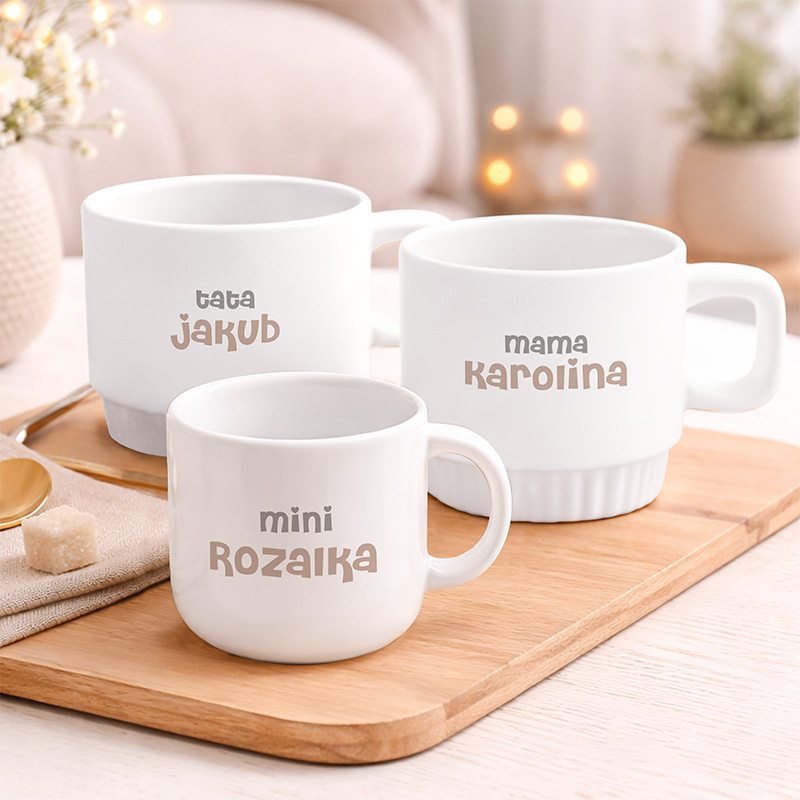 Białe filiżanki z napisem „tata Jakub”, „mama Karolina” oraz „mini Roża” na drewnianej tacy w jasnym wnętrzu.