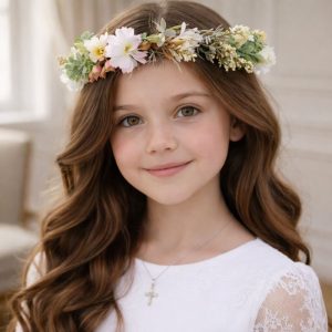 Dziewczynka z długimi, falującymi włosami, nosząca wianek z kwiatów na głowie, uśmiecha się w białej sukience.