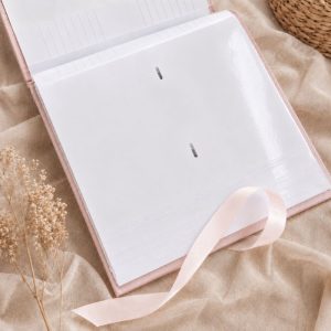 album-welurowy-rozowy-ze-wstazkami Otwarte włoskie białe notes w jasnoróżowej okładce, z widocznymi brzegami stron i wstążką. W tle tkanina i delikatne suche kwiaty.