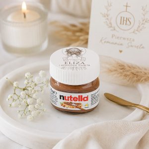 Słoik Nutelli na porcelanowej tacy, obok kwiaty i świeca, w tle widoczna kartka z napisem "Pierwsza Komunia Święta".