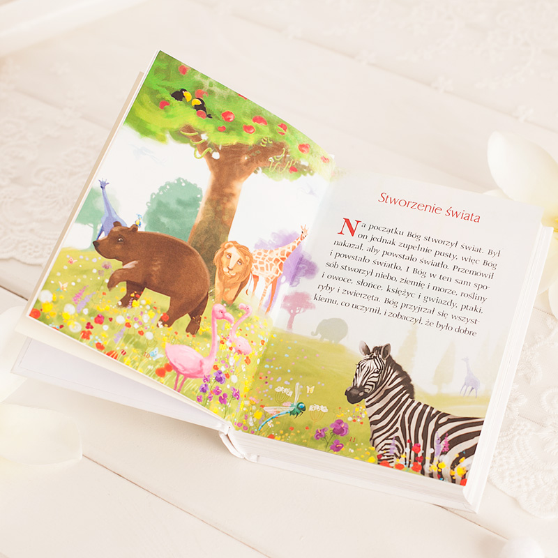 Ilustracja przedstawia otwartą książkę z kolorowymi rysunkami zwierząt w naturalnym środowisku. W centralnej części widoczny jest miś, zebra, żyrafa oraz flamingi, otoczone kwiatami i drzewami. Na stronie znajduje się tekst dotyczący stworzenia świata.