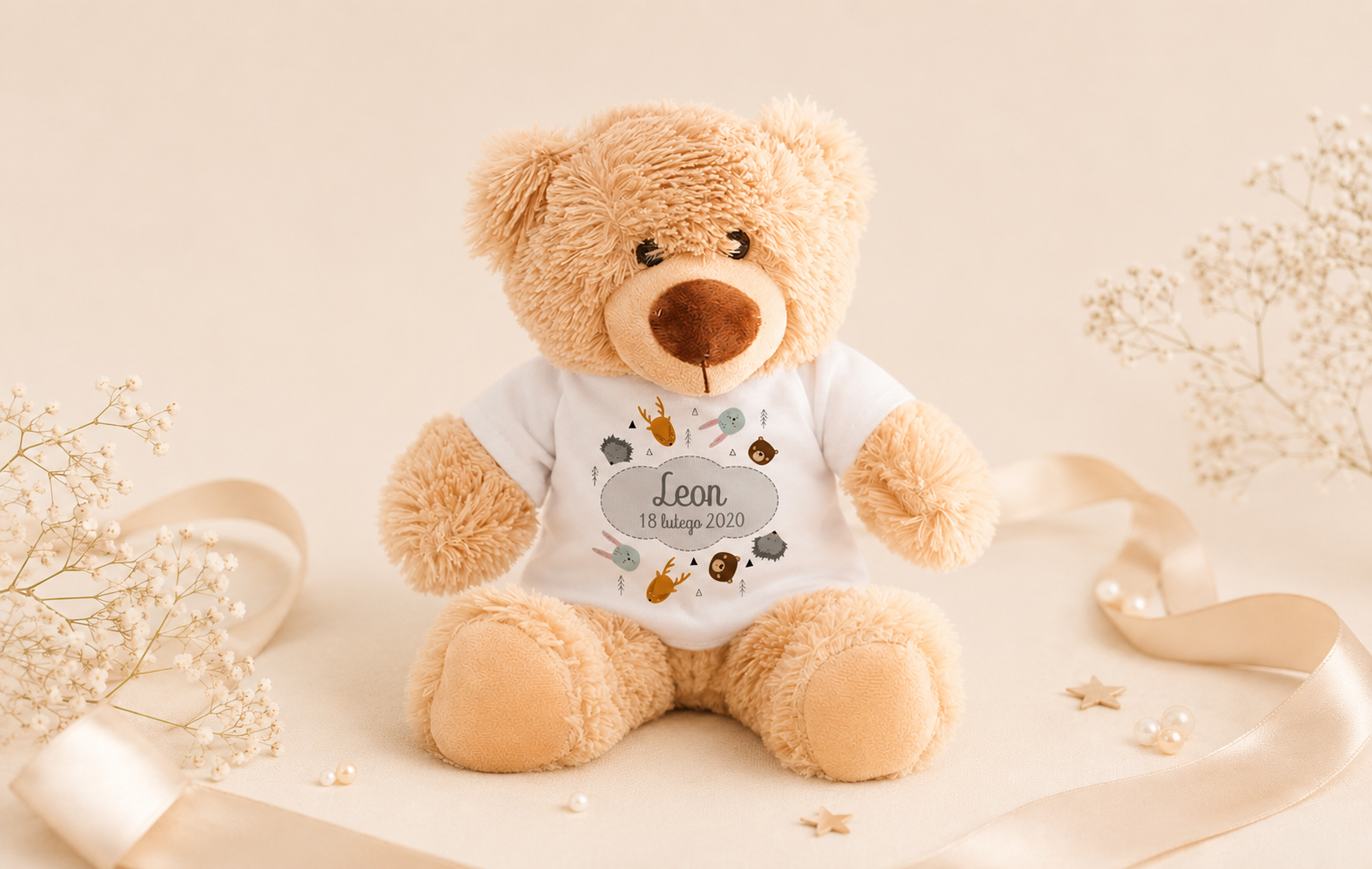 Miś teddy w białej koszulce z napisem "Leon 18 lutego 2020", otoczony delikatnymi kwiatuszkami i złotymi wstążkami.