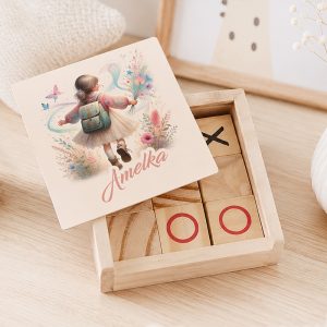 Drewniane pudełko z grą w kółko-krzyżyk, na wieczku ilustracja dziewczynki z plecakiem i imieniem „Amelka”.