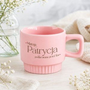 Różowy kubek z napisem, umieszczony na jasnym tle, obok gałązki kwiatów.