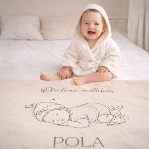 personalizowany-recznik-dla-maluszka.jpg Uśmiechnięte dziecko w jasnym szlafroku siedzi na łóżku z rysunkiem śpiącego niemowlęcia na pościeli. W tle widoczny miękki materiał łóżka.